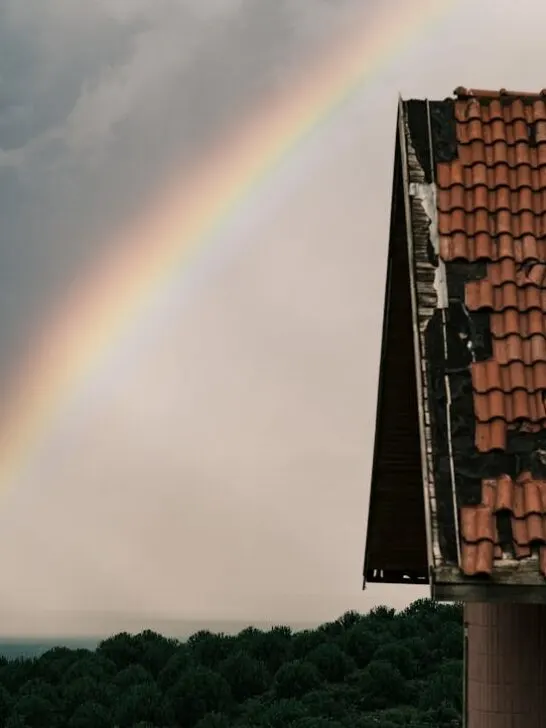 A damaged roof after a storm with a rainbow in the background. Understanding roof warranties is essential as a homeowner to protect your investment.