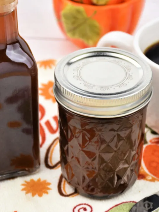 Two bottles of Homemade Pumpkin Spice Syrup sitting next to a cup pf coffee and decorative spoon on a Fall-colored background