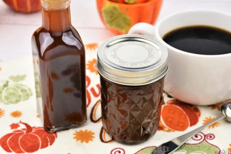 Two bottles of Homemade Pumpkin Spice Syrup sitting next to a cup pf coffee and decorative spoon on a Fall-colored background