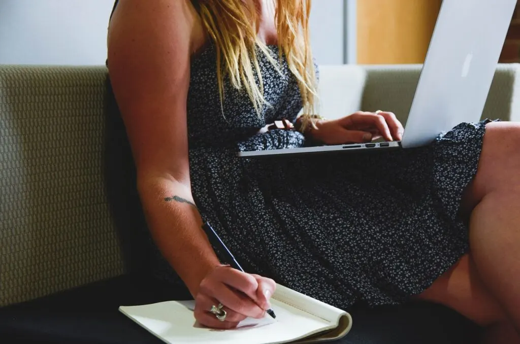 A woman sitting on the couch with her laptop and taking notes in a notebook