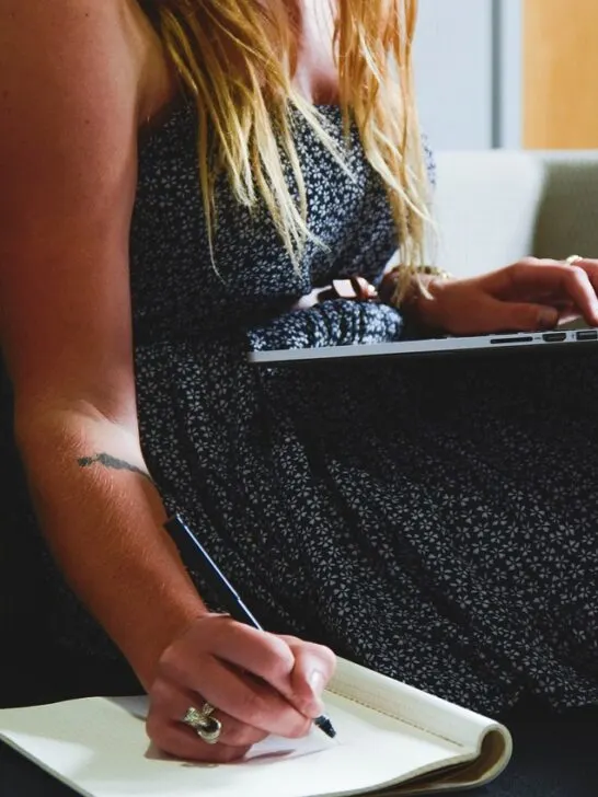 A woman sitting on the couch with her laptop and taking notes in a notebook