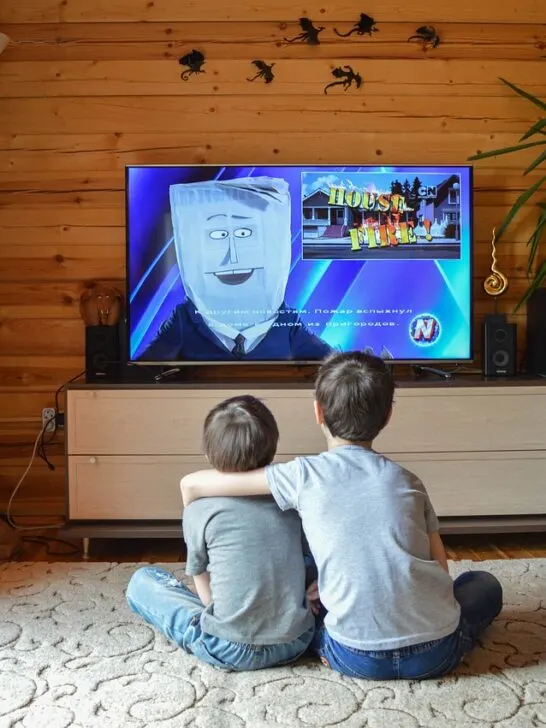 Two kids sitting in front of a television watching a tv that is secured on a tabletop tv stand