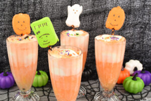 Four Halloween ice cream floats made of orange soda and vanilla ice cream, topped with Halloween Peeps marshmallows on festive straws on black tabletop.