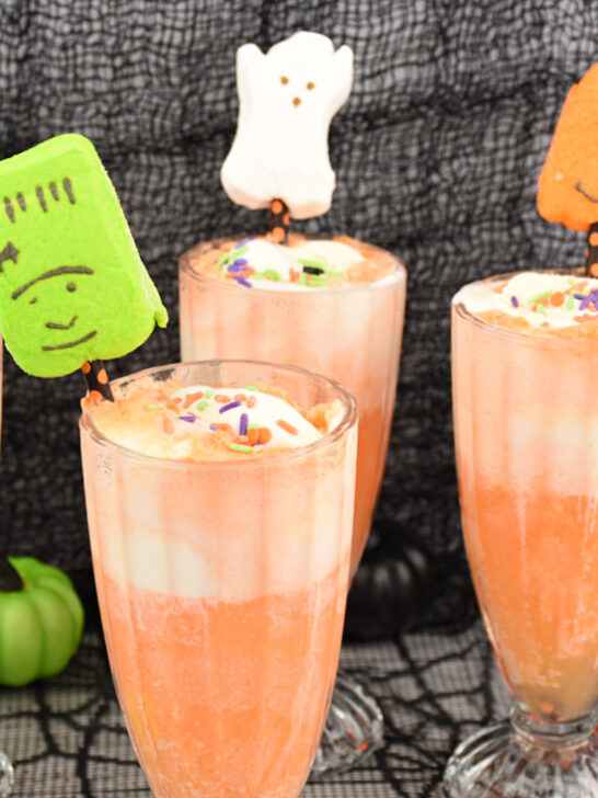 Four Halloween ice cream floats made of orange soda and vanilla ice cream, topped with Halloween Peeps marshmallows on festive straws on black tabletop.