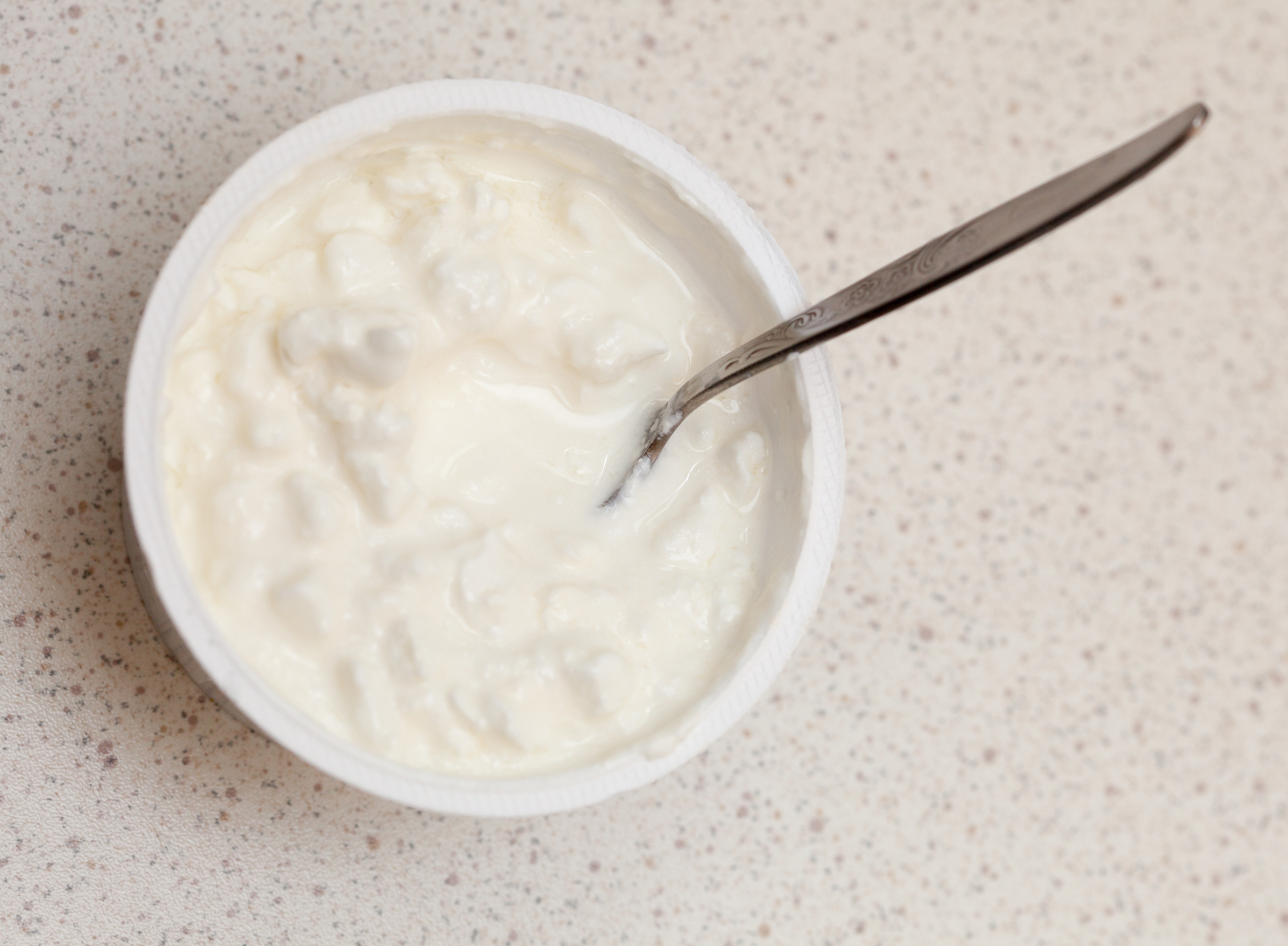 Close-up of white cottage cheese in plastic container and teaspoon on kitchen table background. Food, diet and healthy nutrition concept. Studio shot.