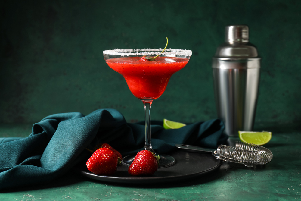Glass of strawberry daiquiri cocktail, shaker and strainer on color background