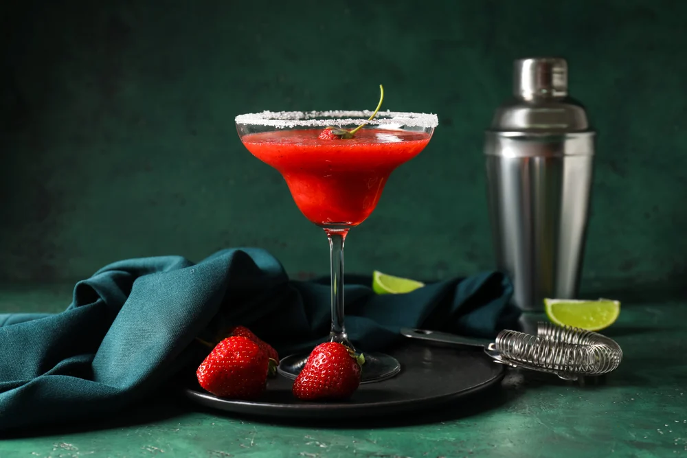 Glass of strawberry daiquiri cocktail, shaker and strainer on color background