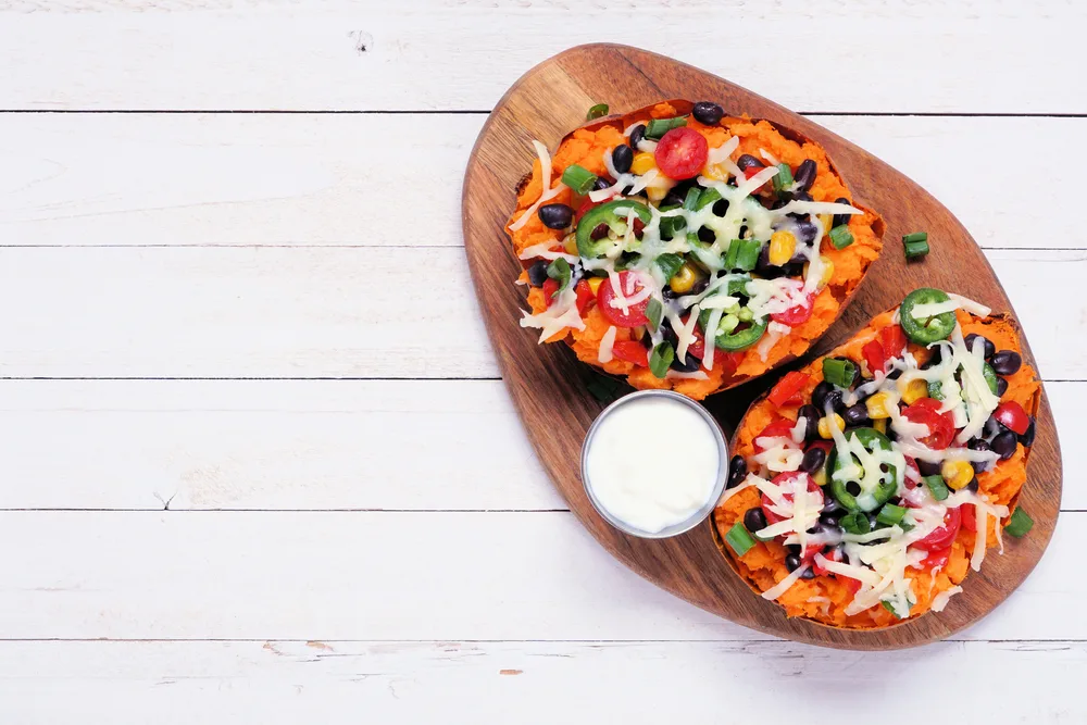 Baked sweet potato tacos stuffed with black beans, vegetables and cheese. Above view on a white wood background.