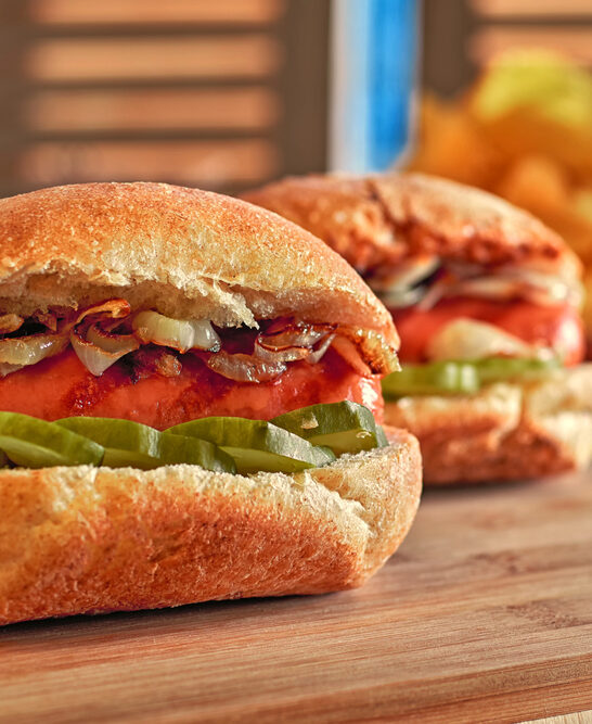 Hot dog with fried onions and marinated cucumbers. Potato chips. Classic fast food on wooden table. Warm glow. — Stock Photo, Image