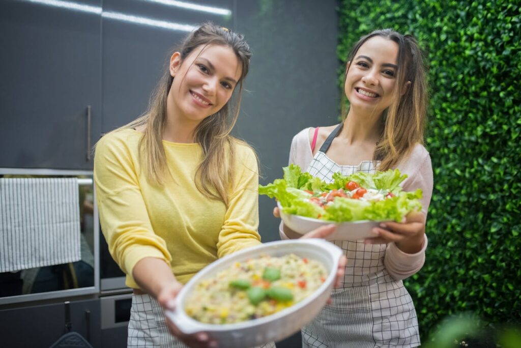 Two women holding out fresh vegetables and a plate of healthy food. Fresh vegetables are part of a Balanced Diet Reduces Stress and Anxiety.