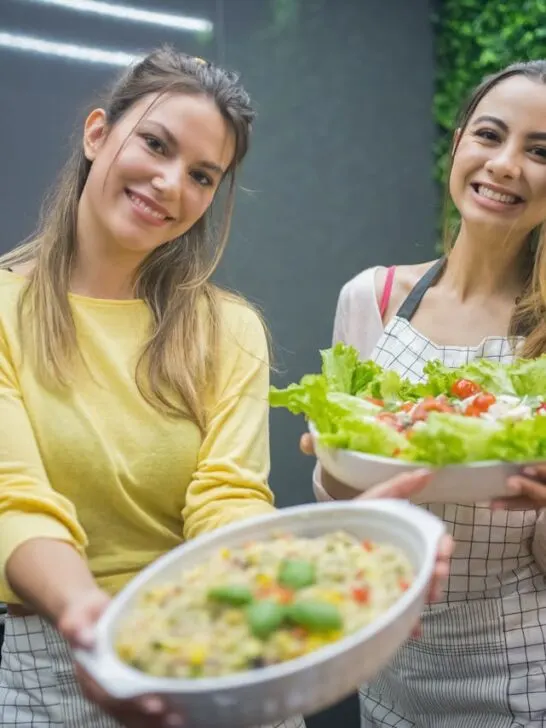 Two women holding out fresh vegetables and a plate of healthy food. Fresh vegetables are part of a Balanced Diet Reduces Stress and Anxiety.