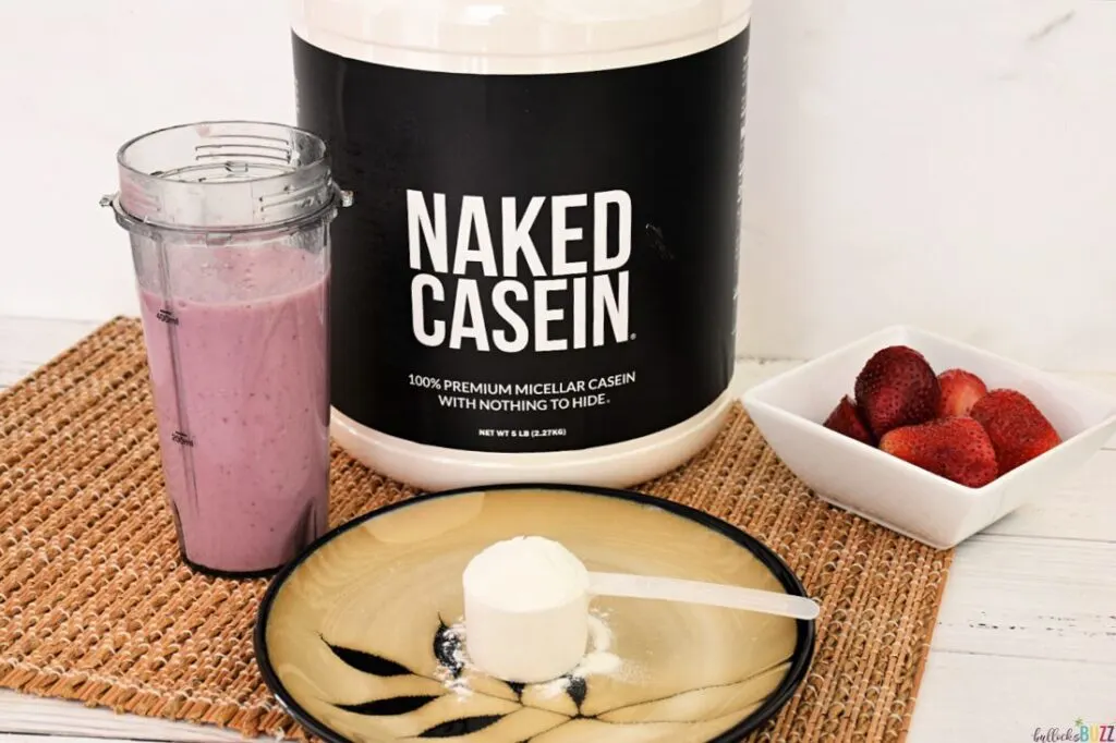 A container of Naked Casein next to a strawberry smoothie, a plate with a scoop of casein and a bowl of strawberries