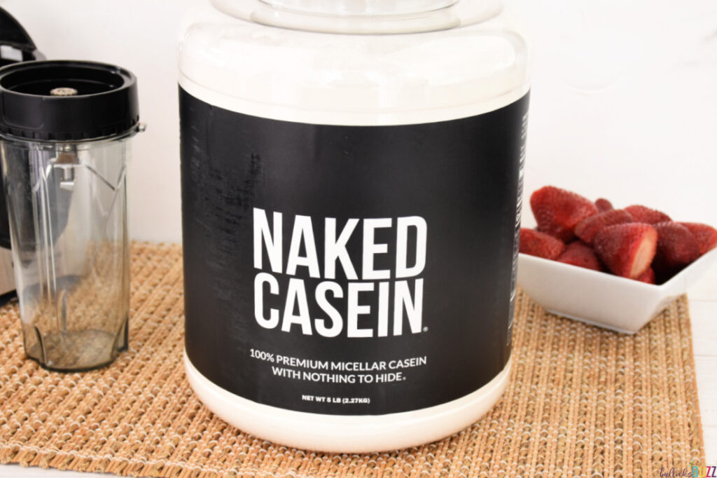 Close up image of a large container of Naked Casein sitting next to a bowl of strawberries and an empty blender cup.