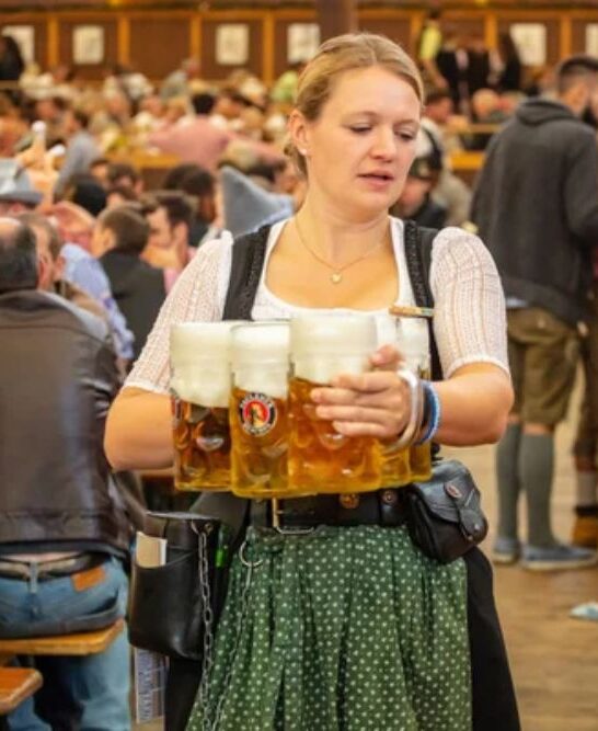 October 7, 2018. Munich, Germany, Oktoberfest, waitress in tyrolean costume holding beers, tent interior background — Photo by gioiak2