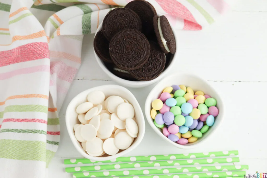 Ingredients needed to make Oreo Flower Pops, including Oreos, pastel M&Ms, and white chocolate melts, are laid out in bowls on a white table