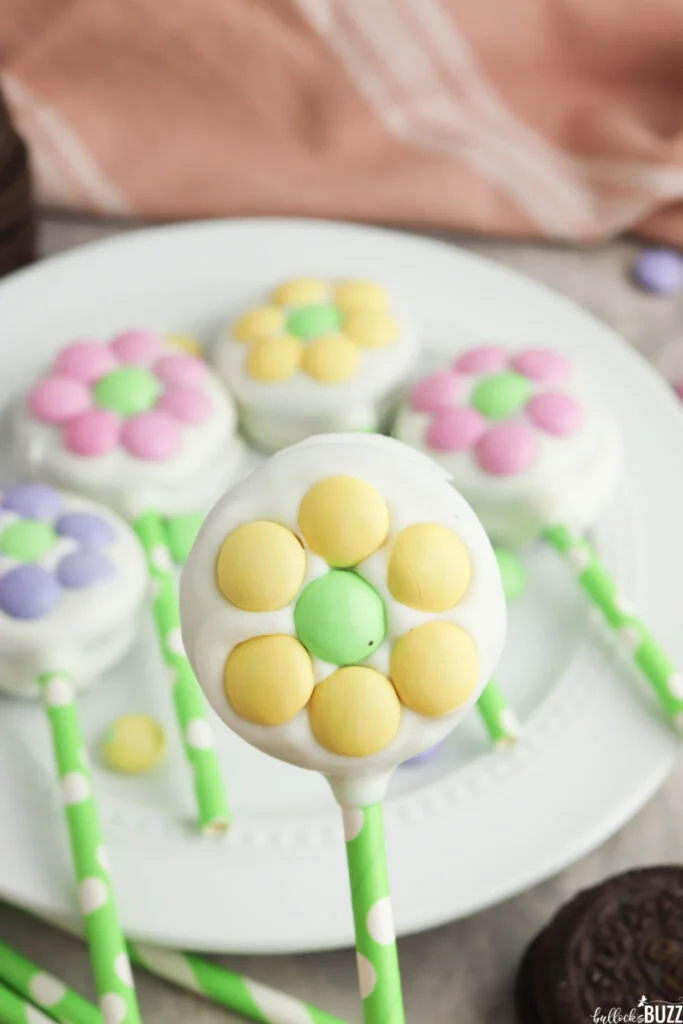 One of the Flower Oreo Pops being held up above a plate filled with of additional Easter flower pops.