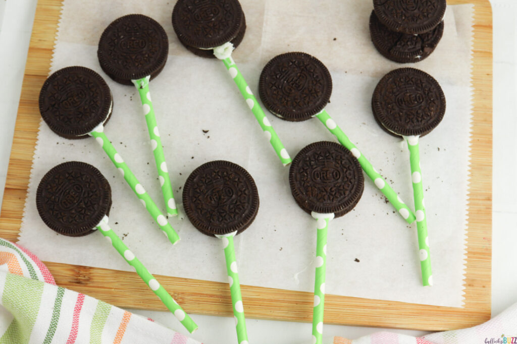 Oreos laid out on parchment paper with straws dipped in the melted chocolate then inserted in between the two cookies