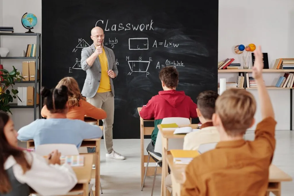 Kids in class facing a blackboard with one raising his hand. Multivitamins for growing children also help with mental and intellectual development,