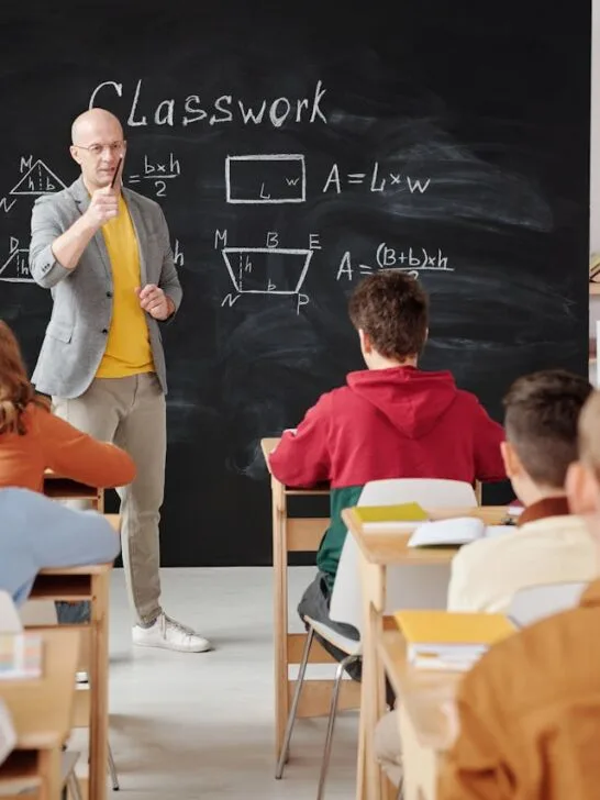 Kids in class facing a blackboard with one raising his hand. Multivitamins for growing children also help with mental and intellectual development,