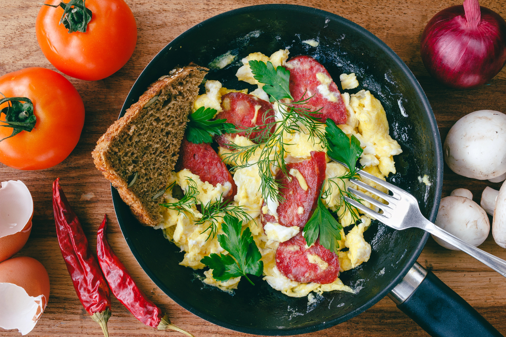Fresh cooked scrambled eggs in pan with sausage and herbs. Bread, fork, vegetables on wooden board, top view. — Stock Photo, Image