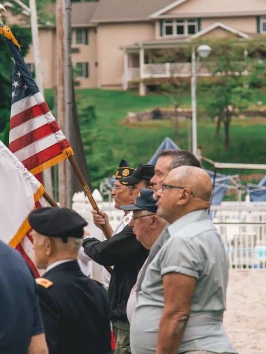 Group of veterans, both young and senior, standing together with one holding the U.S. flag and another holding another military flag, representing unity and support through senior veterans benefits.