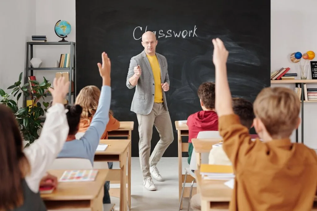 A teacher standing in front of a blackboard with students sitting in their desks raising their hands