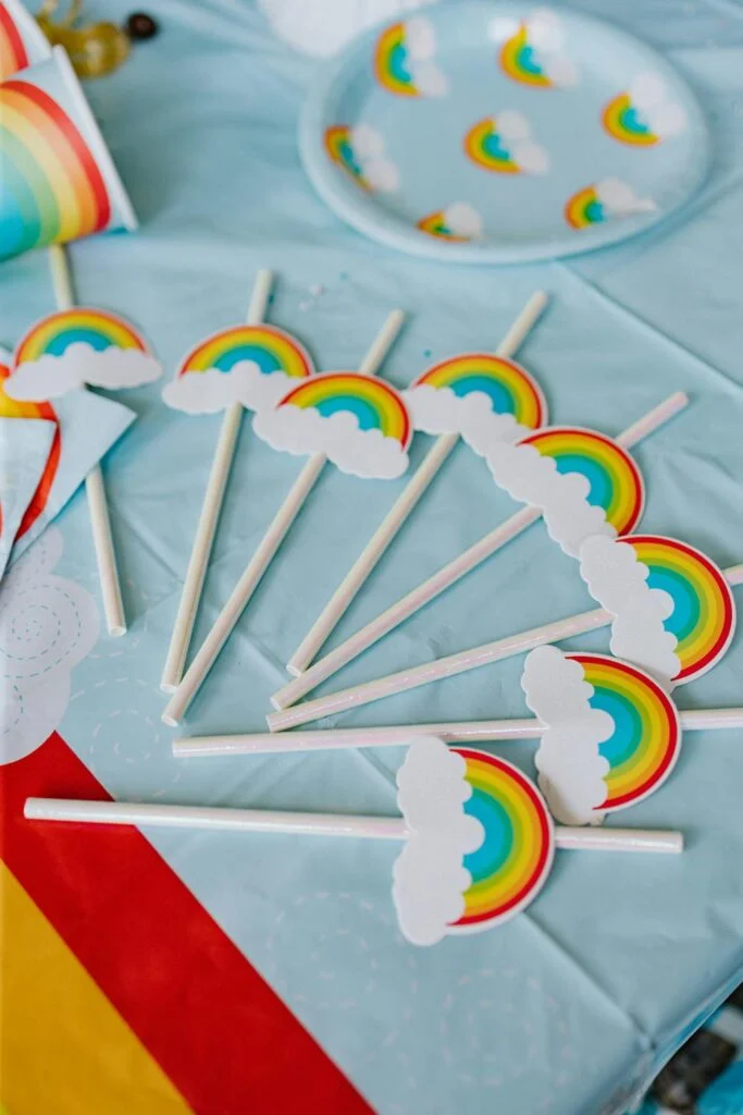 Straws with rainbow toppers laid out on a table with rainbow plates and rainbow cups as part of a rainbow themed summer party for kids