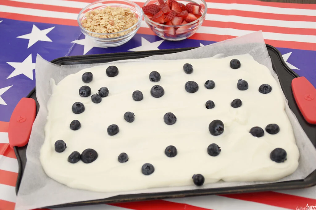 Blueberries are scattered across yogurt spread out on a baking sheet