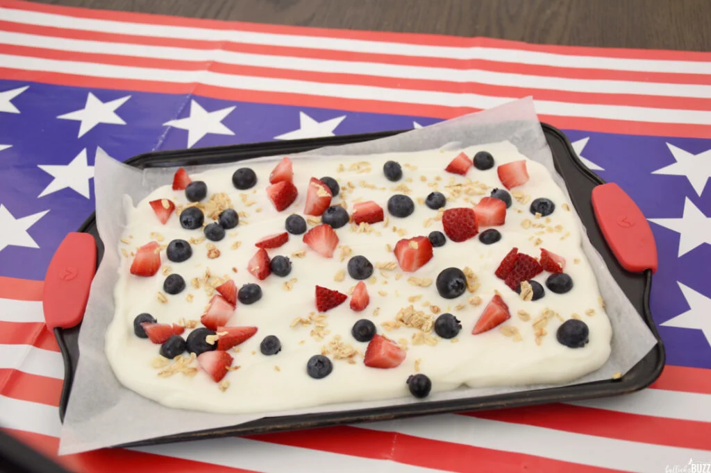 Strawberries and granola are added to the blueberries on top of greek yogurt spread on a baking sheet to make Frozen Yogurt Bark for the 4th of July