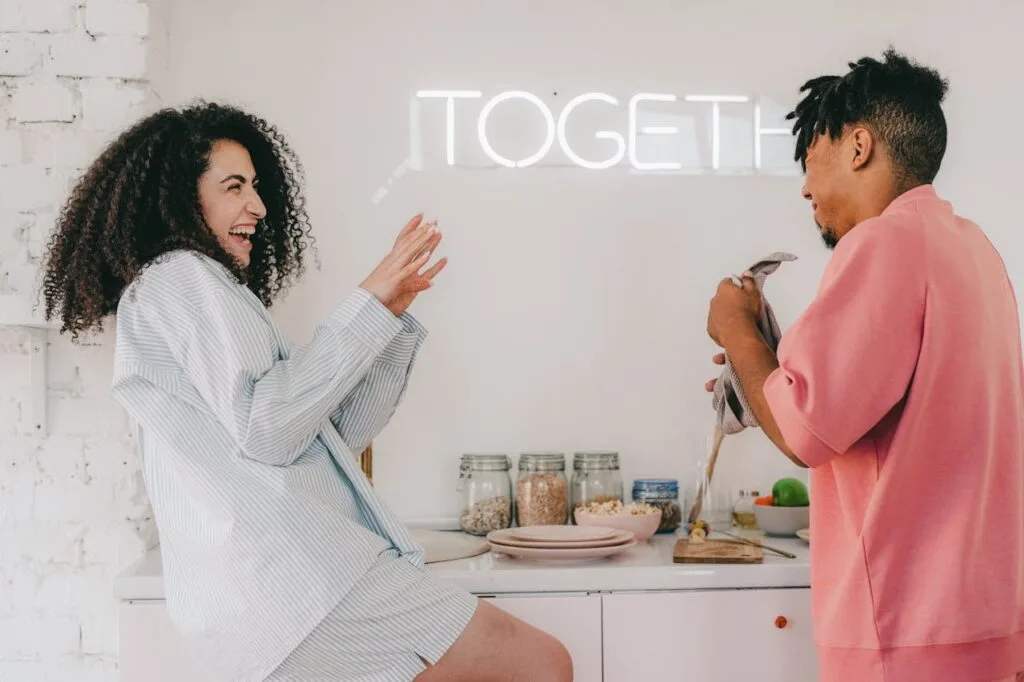 A man and woman laughing in front of a counter with food on it and a neon sign on the wall.