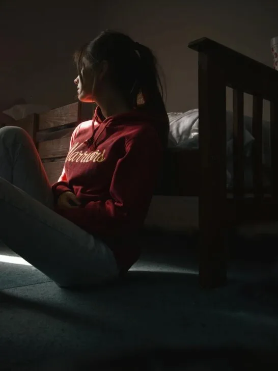 A teen sitting on the floor leaning against her bed and looking out the window. She looks depressed which highlights the need for inpatient mental health treatment for teens