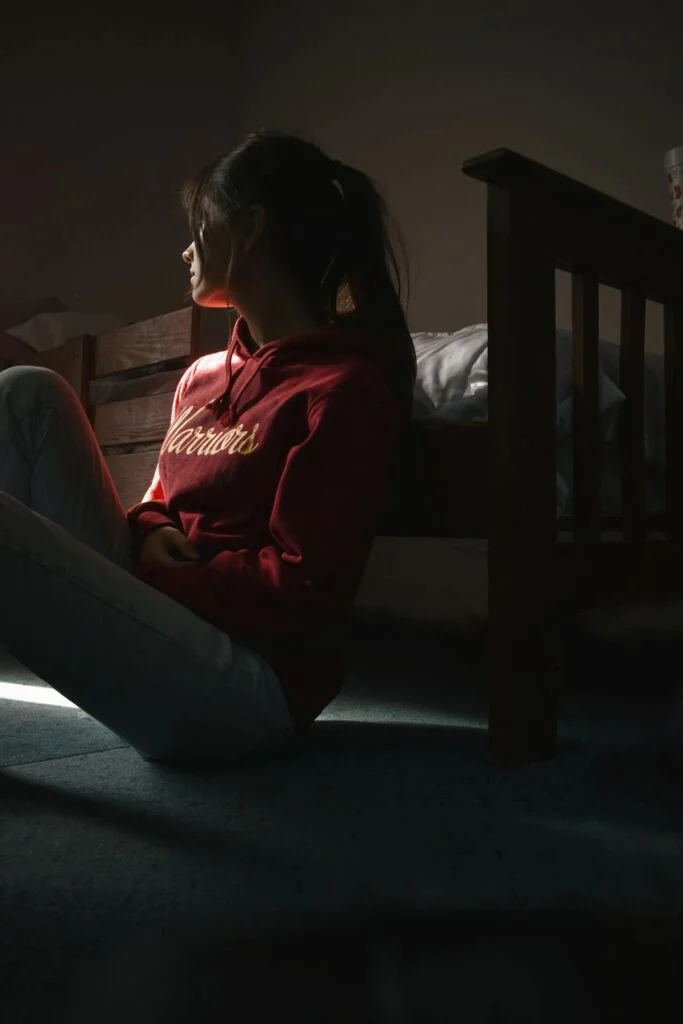 A teen sitting on the floor leaning against her bed and looking out the window. She looks depressed which highlights the need for inpatient mental health treatment for teens