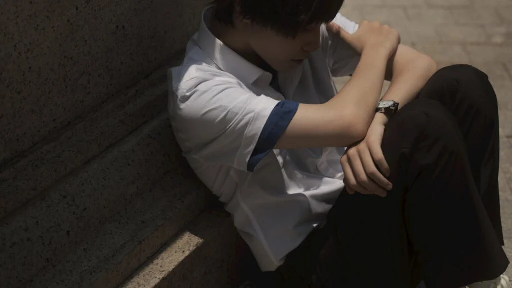 A depressed teenager sitting with his back against a wall looking down at his feet