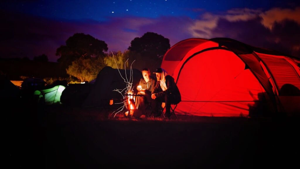 People sitting around a fire with their tents lit up behind them representing several of the most important Family Camping Must-Haves to bring.