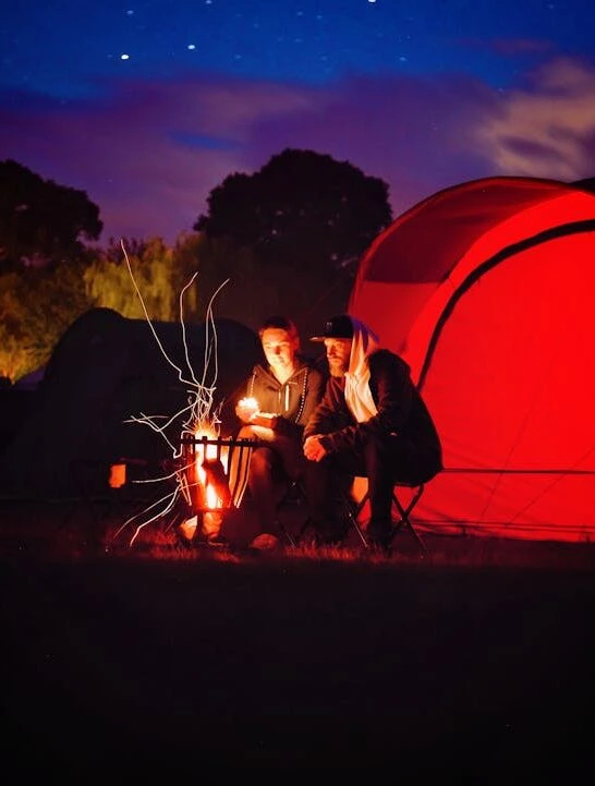 People sitting around a fire with their tents lit up behind them representing several of the most important Family Camping Must-Haves to bring.