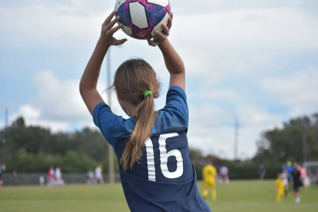 Young girl in a blue soccer jersey holding a soccer ball overhead during a kids’ soccer game