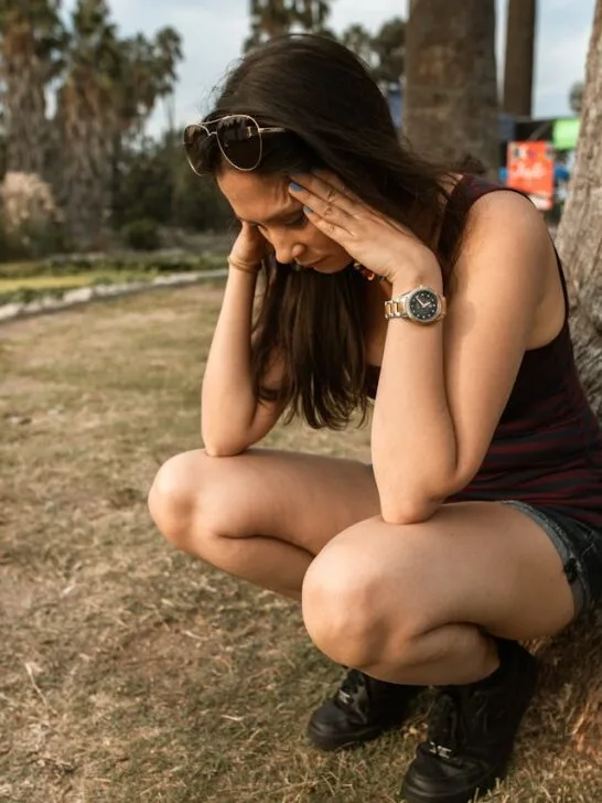 Woman holding her head in stress while sitting outdoors, showing the struggle of how to manage stress and anxiety.