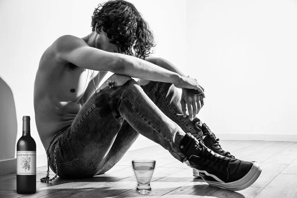 Young man sitting on the floor with head down next to a bottle and glass of alcohol, symbolizing the struggle before taking small steps to sobriety