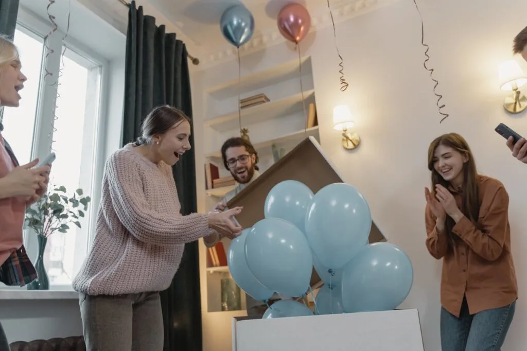 Couple opening a box of balloons during a stress-free gender reveal party, surrounded by smiling friends and family.