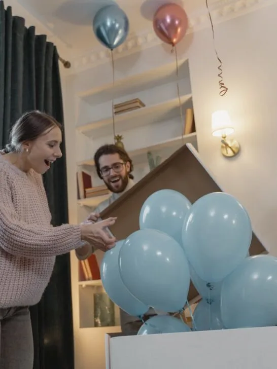Couple opening a box of balloons during a stress-free gender reveal party, surrounded by smiling friends and family.