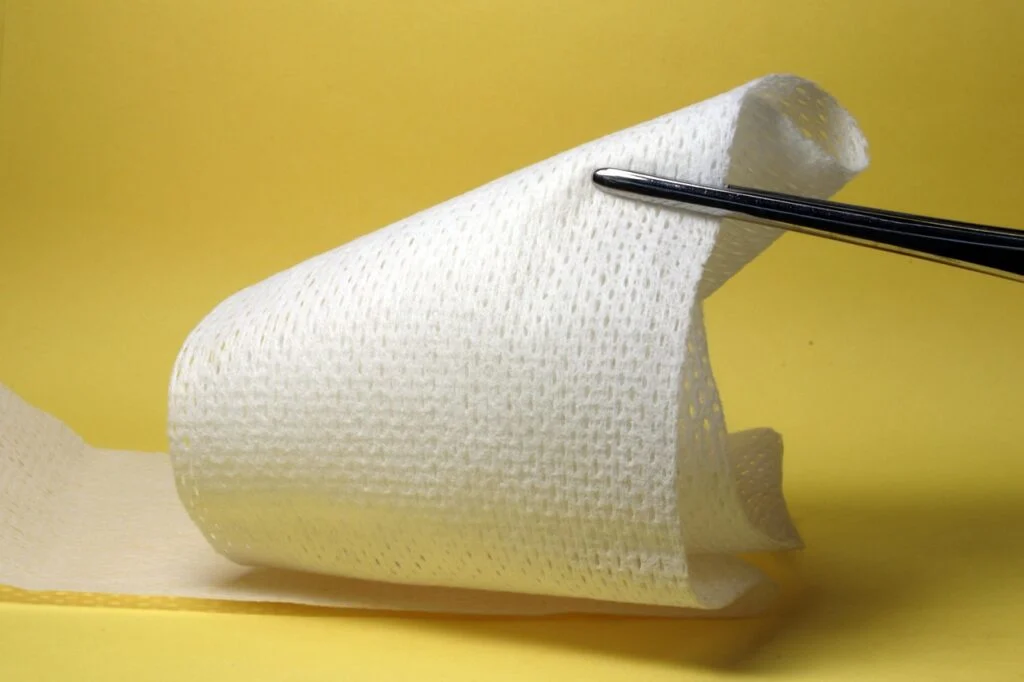 A roll of white gauze bandage being held and measured by metal tweezers against a yellow background, illustrating basic wound care and first‑aid supplies.