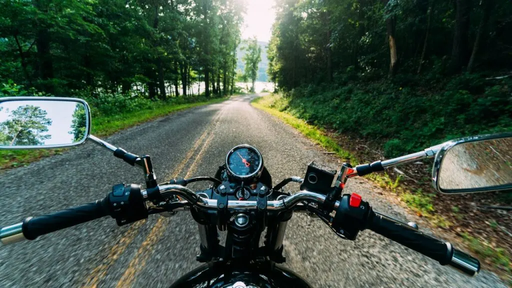 A rider&rsquo;s point of view from the handlebars of a motorcycle cruising down a winding forest road on a sunny day, showcasing the freedom of country riding.