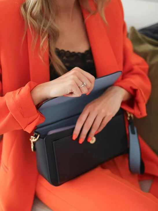Person in a bright orange blazer opening a black designer handbag, showcasing designer handbags worth buying secondhand