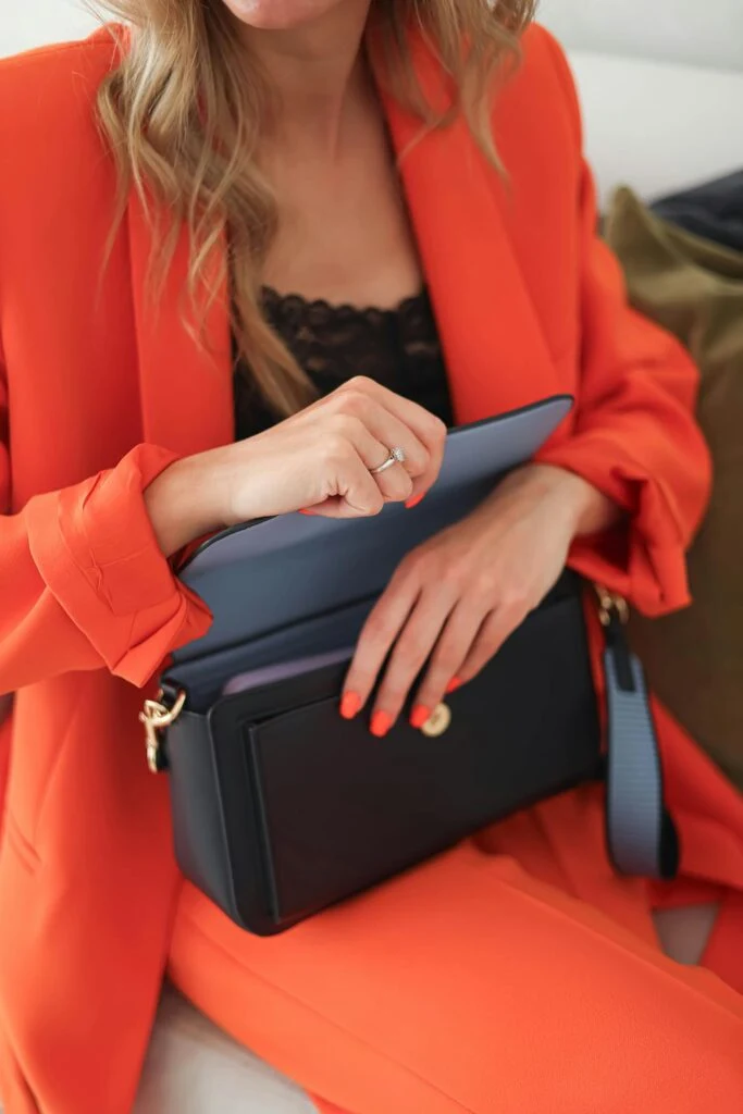 Person in a bright orange blazer opening a black designer handbag, showcasing designer handbags worth buying secondhand