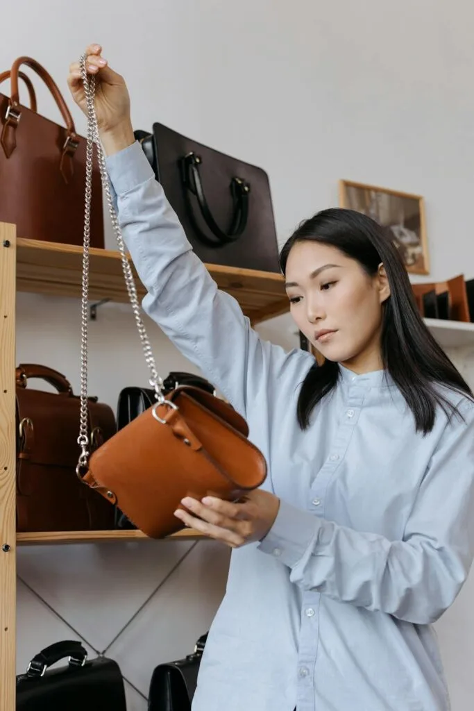 Woman in a store carefully inspecting a brown leather designer handbag with a chain strap among other handbags on display