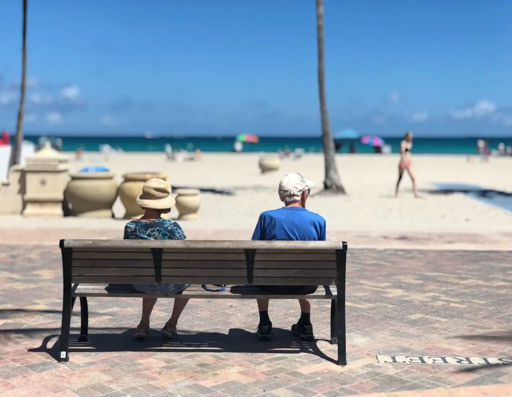Two retired people sitting on a bench facing the ocean, viewed from behind, enjoying a peaceful seaside moment.