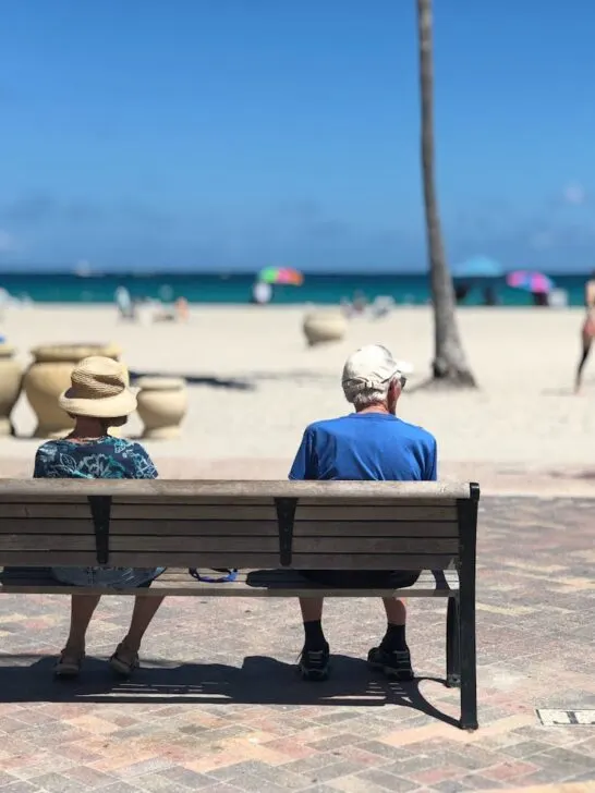 Two retired people sitting on a bench facing the ocean, viewed from behind, enjoying a peaceful seaside moment.