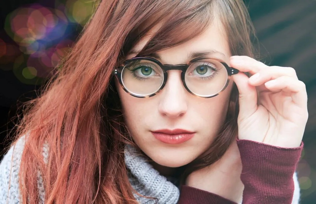 Close-up of a woman adjusting her round prescription eyeglasses, showing clear green eyes and auburn hair.