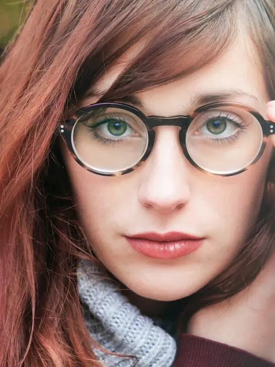 Close-up of a woman adjusting her round prescription eyeglasses, showing clear green eyes and auburn hair.