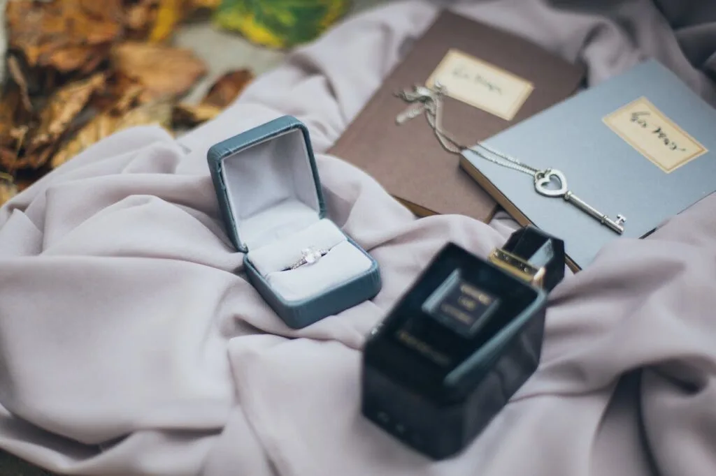 Open ring box with a delicate ring, a heart-shaped key necklace on notebooks, and a perfume bottle arranged on soft fabric with autumn leaves nearby.